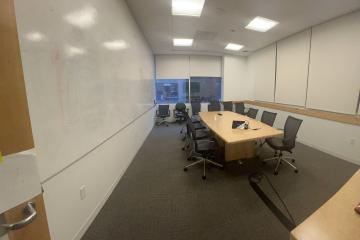 Full view of Pod D conference room from door entry showing grey carpet, whiteboard calls, and central meeting table with chairs.