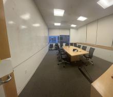 Full view of Pod D conference room from door entry showing grey carpet, whiteboard calls, and central meeting table with chairs.